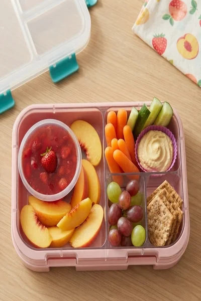 Lonchera rosada con duraznos, fresas, zanahoria, pepino, hummus, uvas y galletas